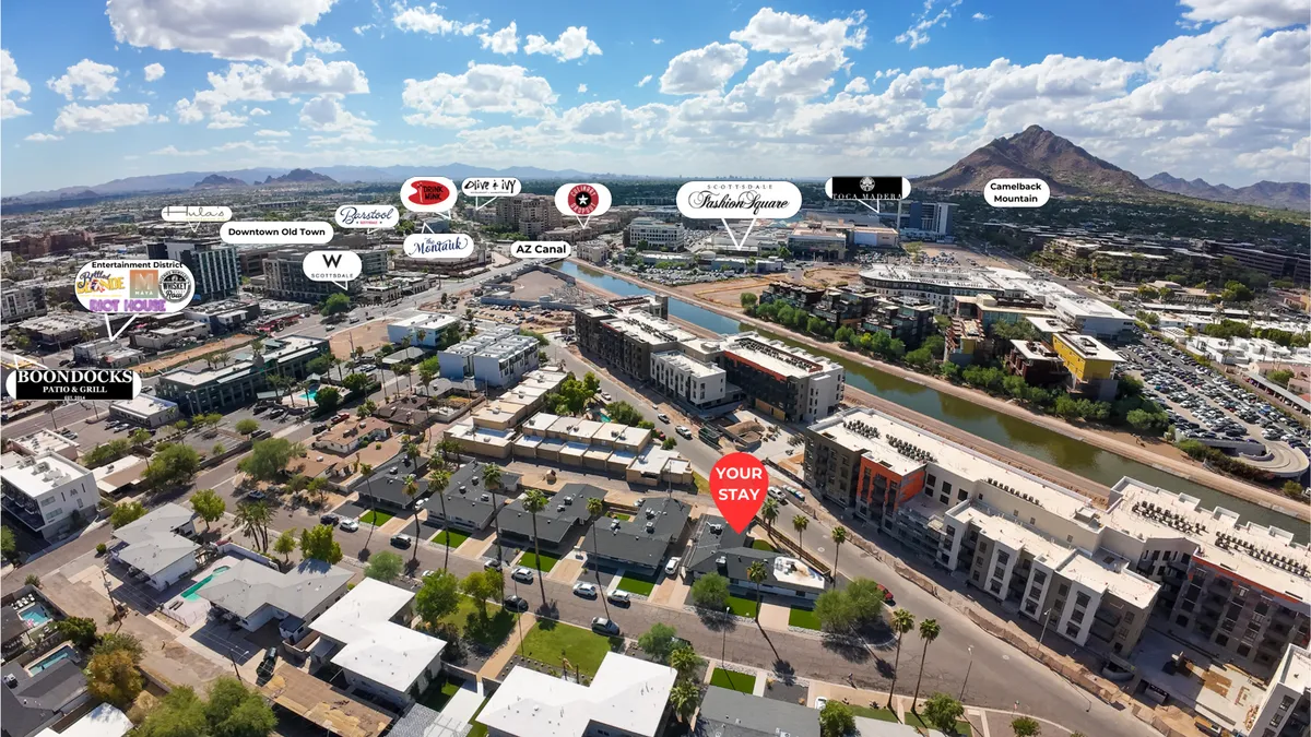 A vibrant view featuring the Arizona Canal winding through Old Town Scottsdale, with Camelback Mountain rising in the background. The image includes the location of 'Your Stay,' nearby buildings, and lively urban activity along the canal.