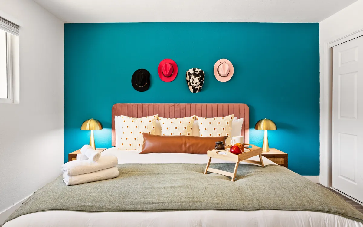 A playful and inviting bedroom scene showing a plush bed adorned with decorative pillows and stylish bedside lamps. A wooden nightstand with a tray, colorful hats on the wall, and soft lighting create a cozy and creative vibe.