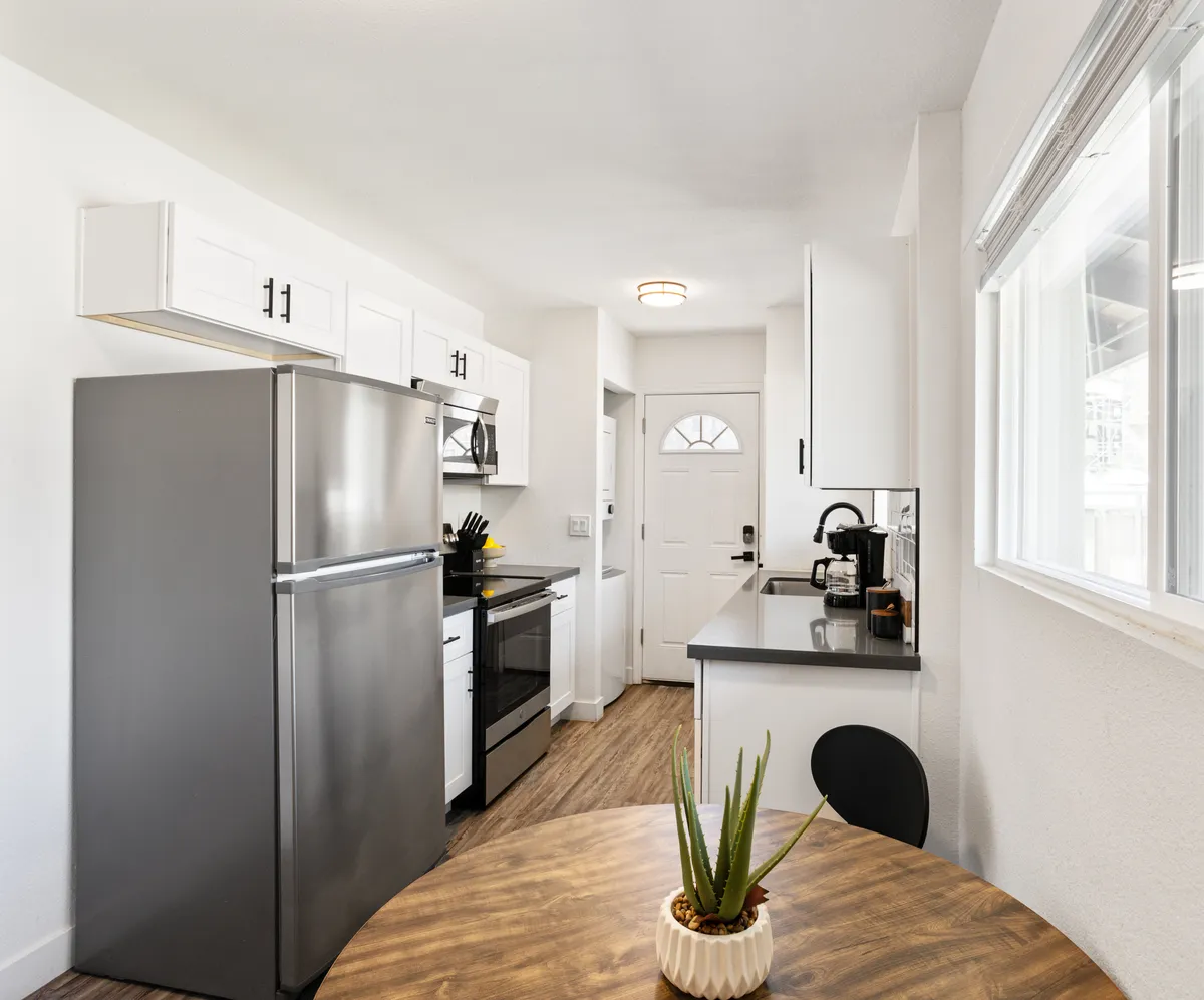A welcoming kitchen scene featuring a stainless steel refrigerator, a handy kitchen island, and a dining table with a green plant as centerpiece. The layout encourages both meal preparation and casual dining in a bright, open setting.