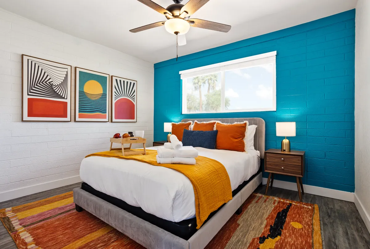 A cheerful bedroom illuminated by natural light, featuring a bed with colorful bedding, artwork on the wall, and a window that lets in sunshine. The room exudes a welcoming, cozy atmosphere perfect for rest and relaxation.