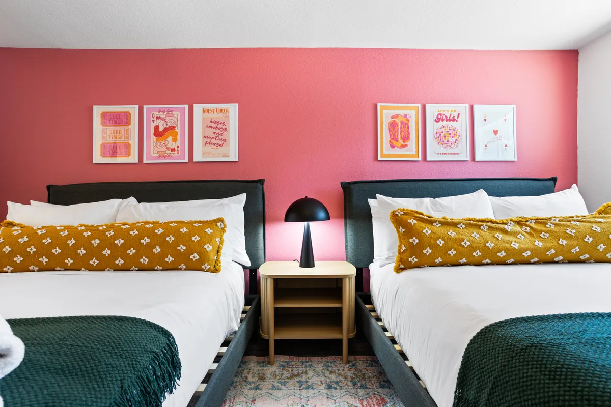 A vibrant, double-bed bedroom with decorative pillows, colorful wall art, a modern bedside lamp, and a textured throw blanket. The space feels playful, perfect for friends or families sharing a room.