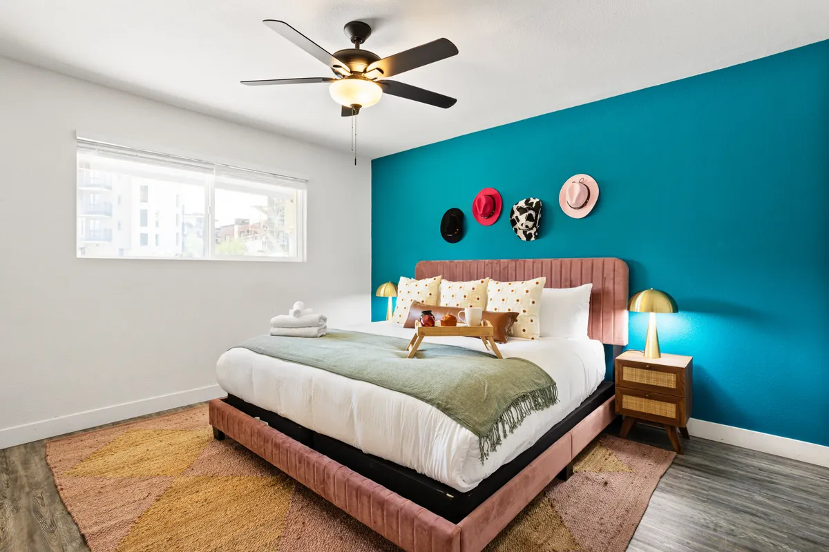 The image showcases a sunlit bedroom featuring a plush bed with a pink velvet headboard, a display of colorful hats on the wall, and a tray with breakfast items invitingly set atop the bedding. The room exudes a cheerful, cozy atmosphere with personal, stylish decor touches.